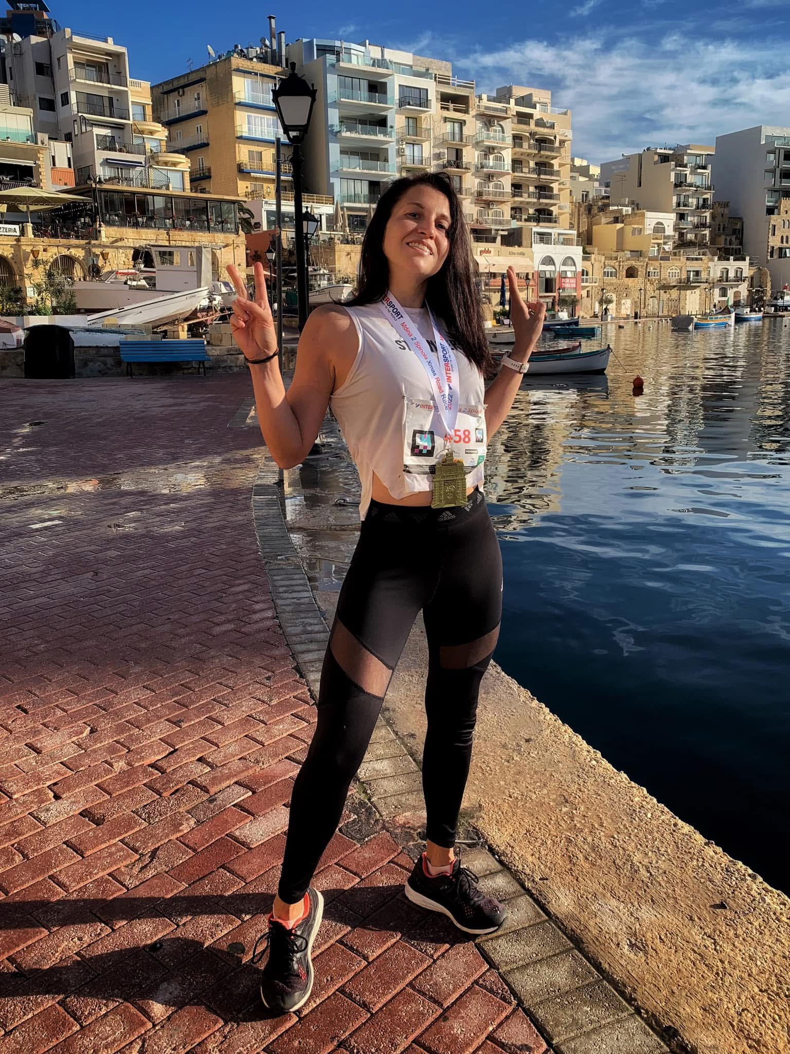Flor celebrating at Spinola Bay after finishing the Mdina to Spinola race, wearing race bib and finisher medal with the harbour and traditional Maltese boats behind her